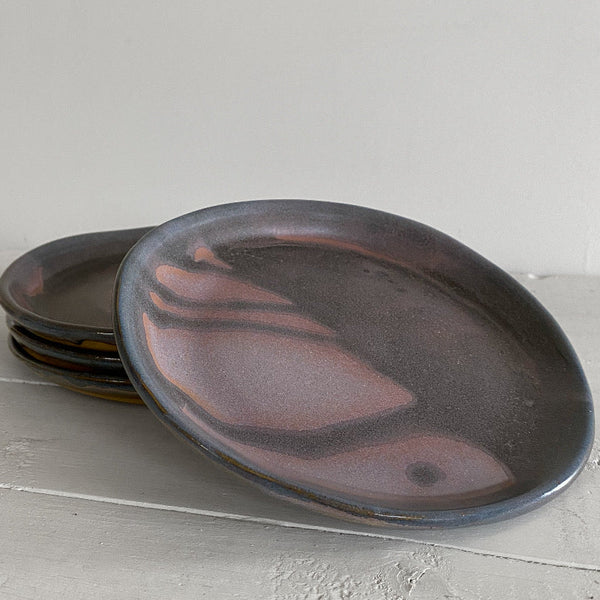 A stack of ceramic plates with a dark charcoal and dusty rose glazed finish, resting on a light gray textured surface against a neutral background.