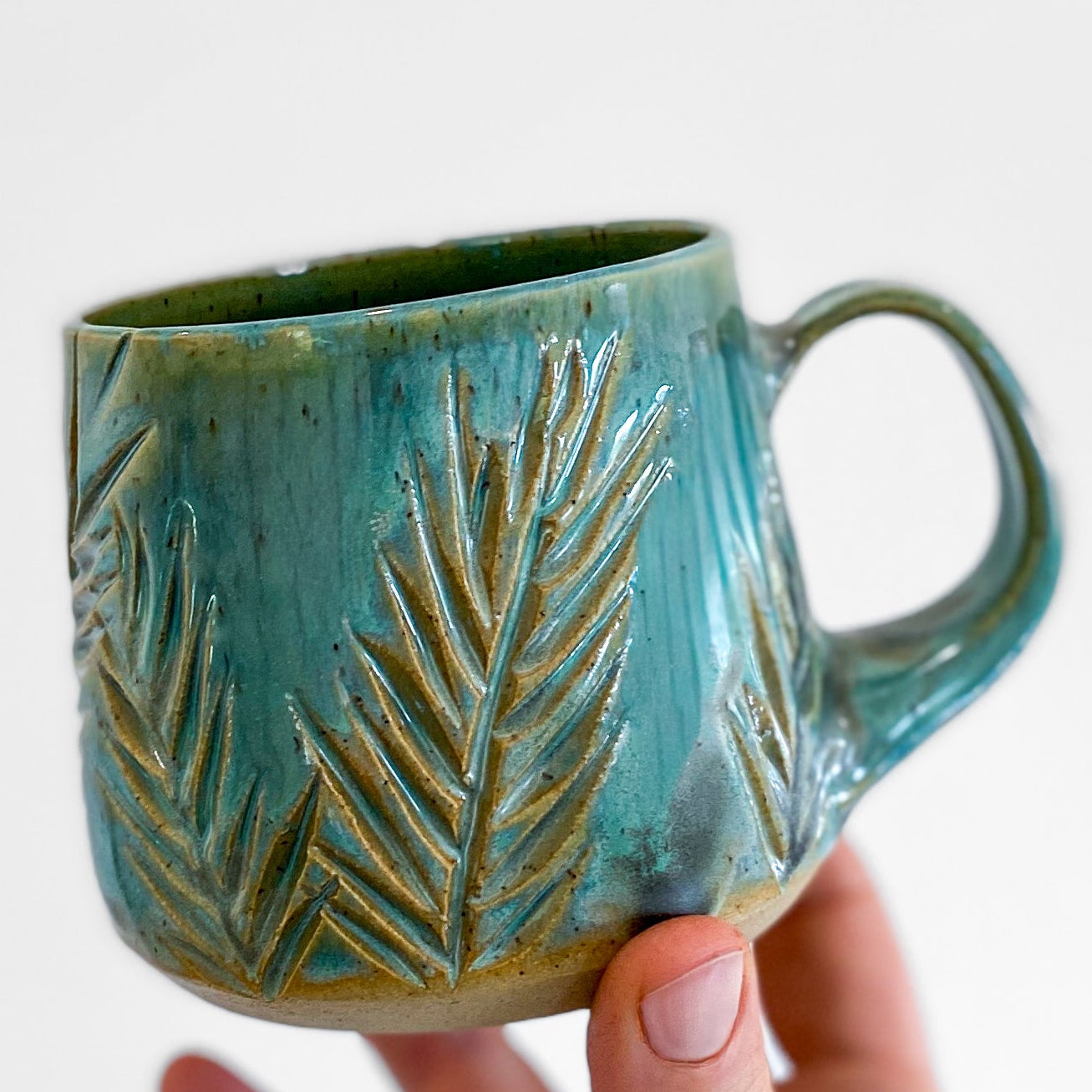 Hand holding a teal ceramic mug with leaf patterns on a light gray background