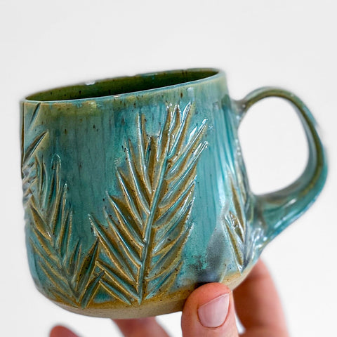 Hand holding a teal ceramic mug with leaf patterns on a light gray background
