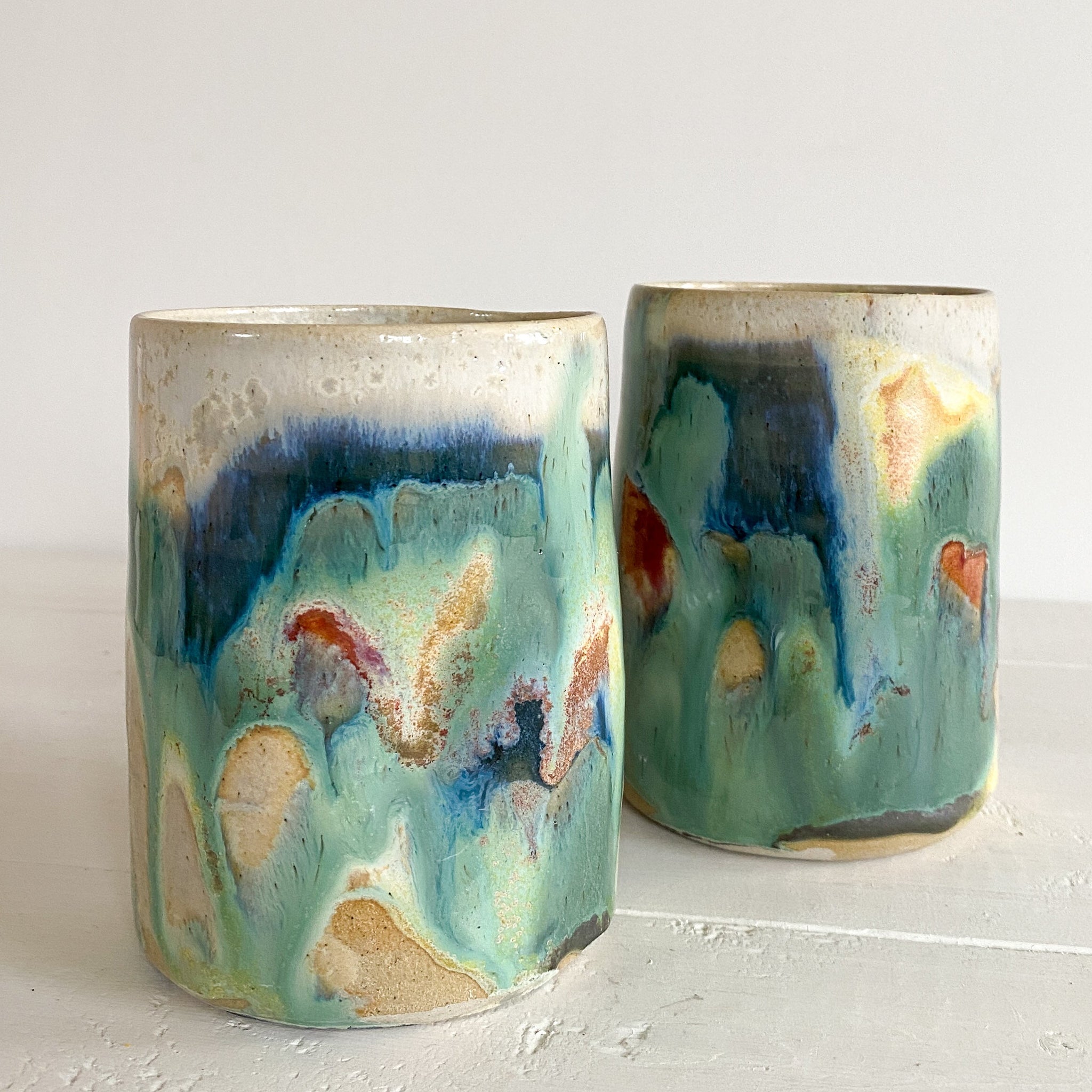Two ceramic tumblers with abstract multicolor glaze on a white surface.
