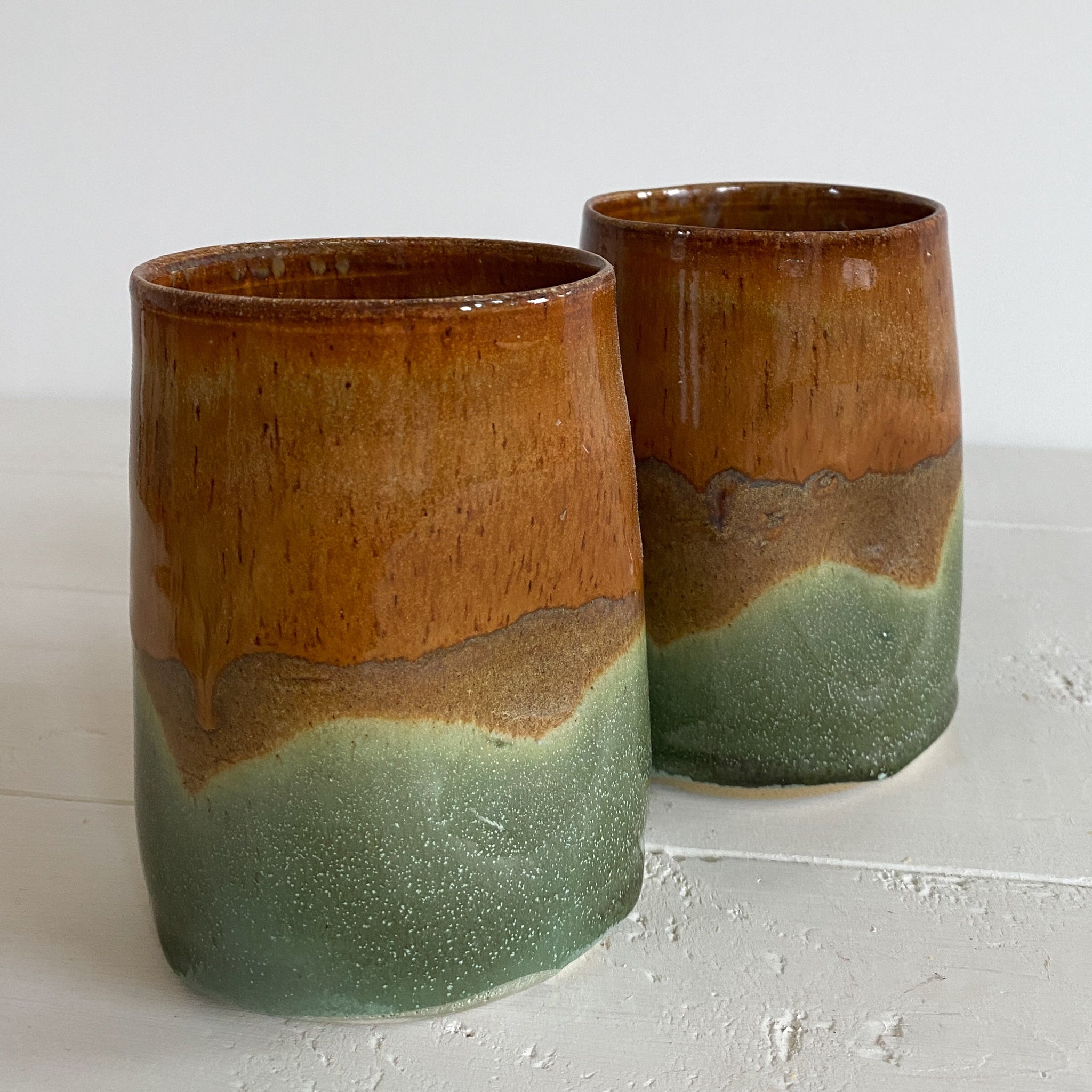 Two ceramic tumblers with a two-tone brown and green glaze on a light gray surface.