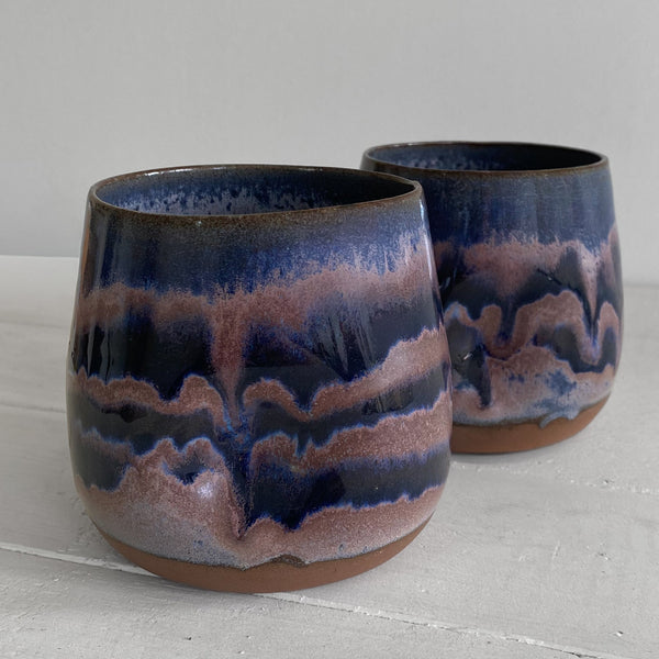 Two handmade ceramic tumblers with a marbled glaze in deep navy blue, purple, and copper-brown, displayed on a light gray surface against a neutral background.