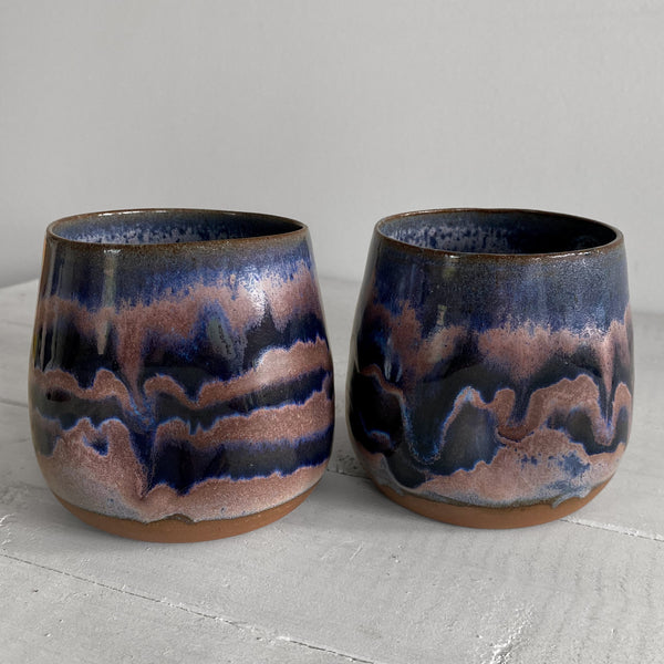 Two handmade ceramic tumblers with a dark blue and copper-brown wavy glaze, displayed side-by-side on a light gray wooden surface against a neutral gray background.