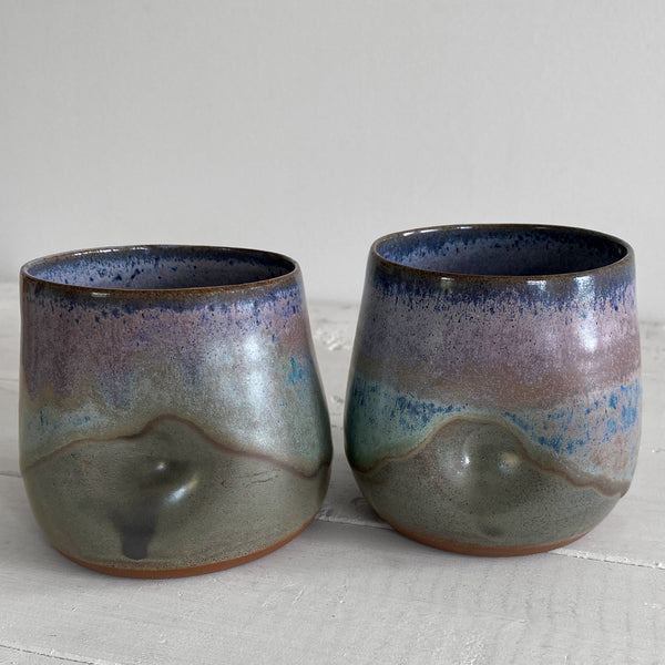 Two handmade ceramic tumblers with a glossy, multi-colored glaze showing shades of blue, purple, green, and brown, set on a white wooden surface against a light gray background.