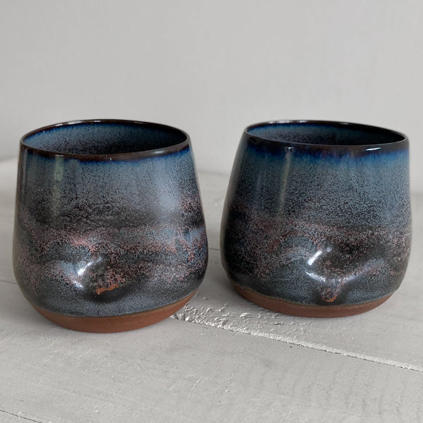 Two handmade ceramic tumblers with a dark blue and black speckled glaze and a terracotta brown base, displayed on a light gray wooden surface.