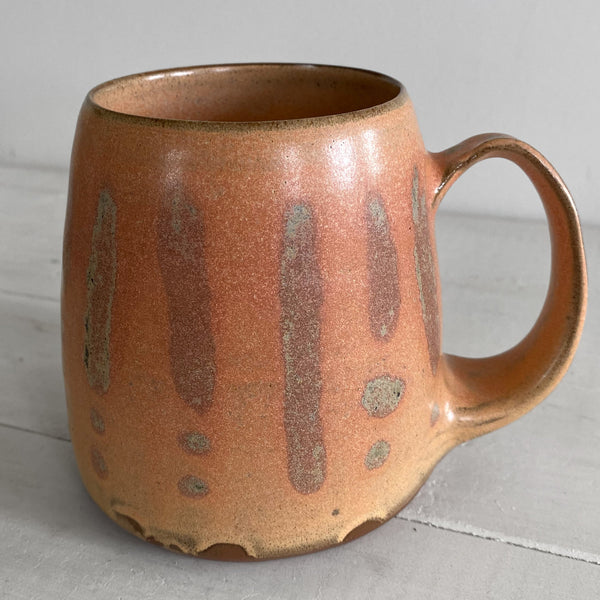 A handmade ceramic mug with a rustic, rounded shape, featuring a warm terracotta brown  peach glaze with vertical brown drips and speckled accents, set on a light gray wooden surface against a neutral gray background.