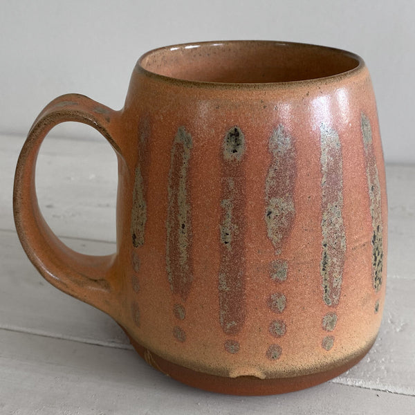 A handmade ceramic mug with a warm terracotta orange-brown glaze, featuring vertical metallic silver stripes and small silver dots on the side, set on a light gray wooden surface against a neutral gray background.