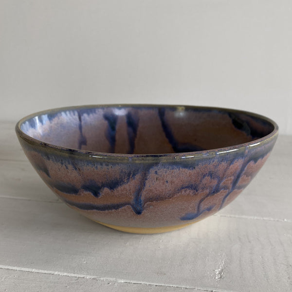 A handmade ceramic bowl with a purple and blue glaze, featuring a dripping pattern, resting on a light gray wooden surface against a neutral background.