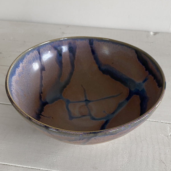 A handmade ceramic bowl with a purple and blue glaze, photographed on a light gray wooden surface against a white wall.